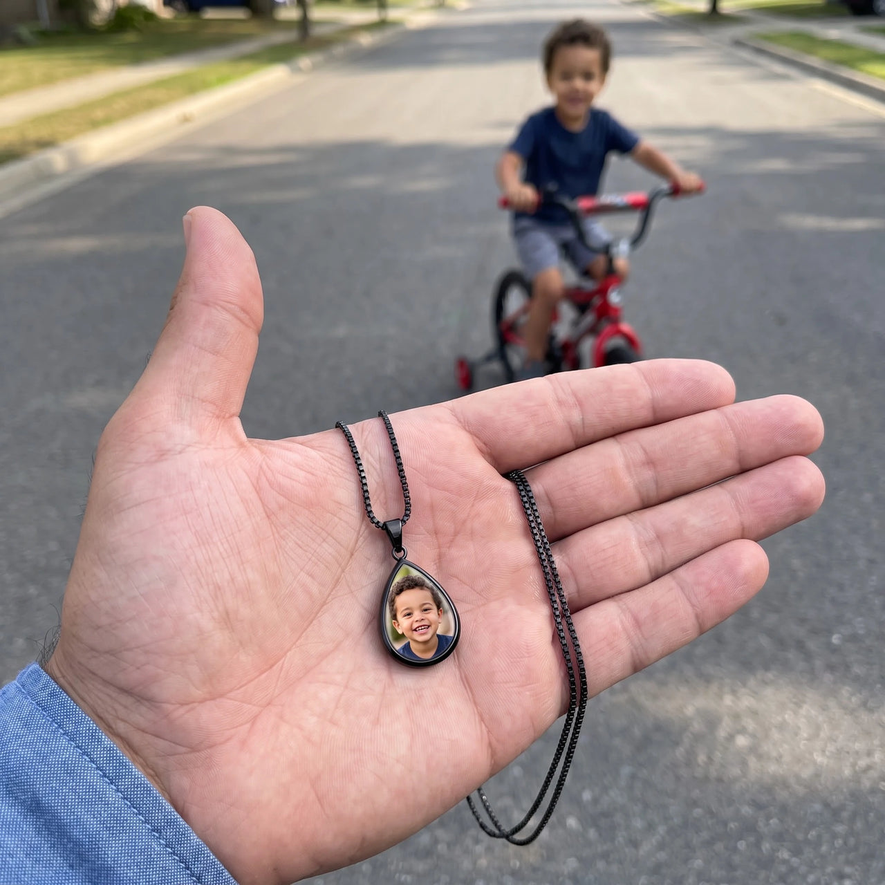 The black teardrop photo pendant in an open palm, real small size, a little boy riding his bike behind | SKU: TPH-DRPN-BLACK